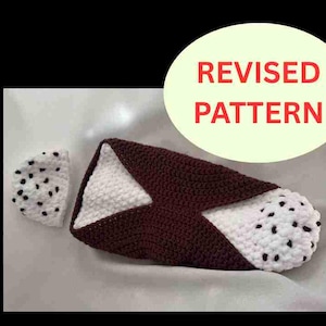 May include: A crocheted cannoli with a brown shell, white filling, and black dot accents. A small, separate crocheted piece with black dots is also present. The image includes a circular graphic with the words "REVISED PATTERN".