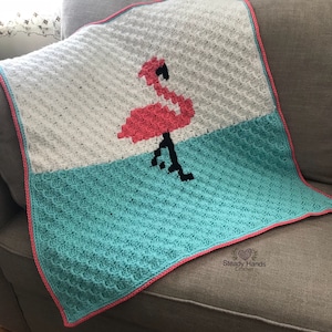 May include: A handmade crochet blanket featuring a pink flamingo design. The blanket is white and turquoise with a pink border. The flamingo is pink with black legs and beak. The blanket is draped over a gray couch.