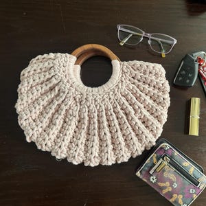 May include: A beige crochet handbag featuring a semi-circular shape with a ribbed texture and two rounded wooden handles. The bag is accompanied by a pair of clear-framed glasses, a black car key fob, a gold lipstick tube, and a patterned wallet.