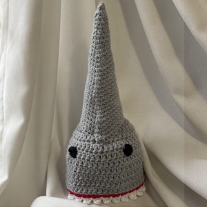 May include: A gray crocheted shark hat with a long, pointed top. The hat features black eyes and a red and white trim at the base, resembling shark teeth. This handmade hat is a fun accessory.