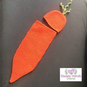 May include: A crocheted carrot baby outfit with a hat and a long body. The outfit is orange with green leaves on the top of the hat. The outfit is made by Steady Hands Crochet.