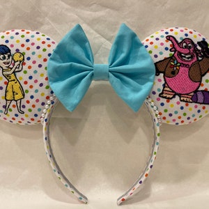 May include: A pair of white mouse ears with a blue bow. The ears are covered in a white fabric with colourful polka dots. One ear features an embroidered image of Joy from the movie Inside Out, and the other ear features an embroidered image of Bing Bong from the movie Inside Out.