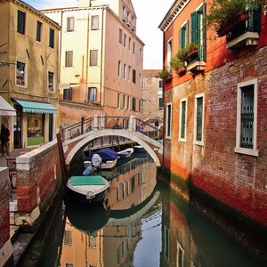 Peut inclure: Un canal étroit à Venise, en Italie, avec un bâtiment en briques à droite et un bâtiment jaune à gauche. Un petit pont traverse le canal, et deux bateaux sont amarrés dans l'eau. L'eau reflète les bâtiments et le ciel.