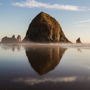 May include: A large, dark rock formation, known as Haystack Rock, rises from the sandy beach. The rock is reflected in the calm, still water, creating a mirror image. The sky is a pale blue with wispy clouds.