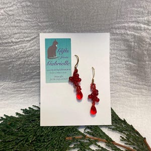 May include: Red beaded dangle earrings with a teardrop-shaped red glass bead at the bottom. The earrings are on a white card with the text "Gifts from Gabrielle" and a cat illustration.