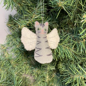May include: A handmade felt cat ornament with angel wings, hanging from a white string. The cat is gray with dark gray stripes, pink ears, and green eyes. The wings are white. The ornament is displayed against a backdrop of green Christmas tree branches.
