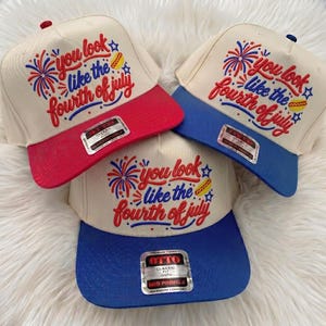 May include: Three baseball caps with off-white crowns and embroidered text that reads you look like the fourth of july. The designs include red and blue fireworks and a hot dog icon. One cap has a red brim, while two have blue brims.