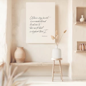 Puede incluir: Una pared beige con una impresi&oacute;n en lienzo blanco con una cita manuscrita en negro: "Cuando mi coraz&oacute;n est&aacute; abrumado, ll&eacute;vame a la roca que es m&aacute;s alta que yo" con el texto "SALMO 61:2" debajo. La impresi&oacute;n est&aacute; colgada en una pared sobre un estante de madera con un jarr&oacute;n beige en &eacute;l. Hay un jarr&oacute;n blanco con flores secas en un taburete de madera.