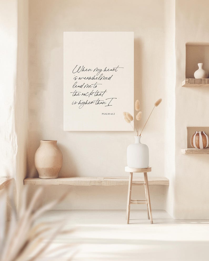 Puede incluir: Una pared beige con una impresi&oacute;n en lienzo blanco con una cita manuscrita en negro: "Cuando mi coraz&oacute;n est&aacute; abrumado, ll&eacute;vame a la roca que es m&aacute;s alta que yo" con el texto "SALMO 61:2" debajo. La impresi&oacute;n est&aacute; colgada en una pared sobre un estante de madera con un jarr&oacute;n beige en &eacute;l. Hay un jarr&oacute;n blanco con flores secas en un taburete de madera.