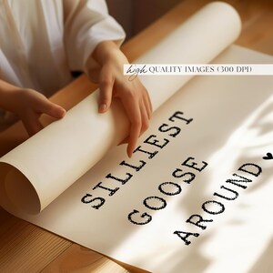 Pu&ograve; includere: Un poster bianco con testo nero che recita "Silliest Goose Around" con un simbolo di cuore alla fine. Il poster viene arrotolato da una mano.