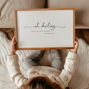 以下が含まれることがあります： 「oh darling don't you ever grow up.」というテキストが書かれた額装されたプリント。白いセーターを着た子供がプリントを持っています。