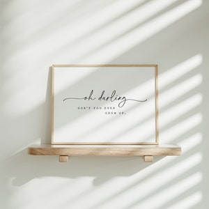 以下が含まれることがあります： 「oh darling don't you ever grow up.」というテキストが書かれた額装されたプリント。プリントは白い壁に面した明るい木製の棚の上に置かれています。