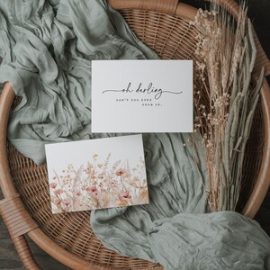 以下が含まれることがあります： 「oh darling don't you ever grow up.」と書かれた白いカード。ピンクと黄色の野花の水彩画が描かれた2枚目のカード。