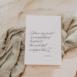 Puede incluir: Una tarjeta blanca con texto en caligraf&iacute;a negra que dice "Cuando mi coraz&oacute;n est&aacute; abrumado, gu&iacute;ame a la roca que es m&aacute;s alta que yo. Salmo 61:2". La tarjeta est&aacute; sobre una superficie de madera con una tela marr&oacute;n claro drapeada sobre ella.