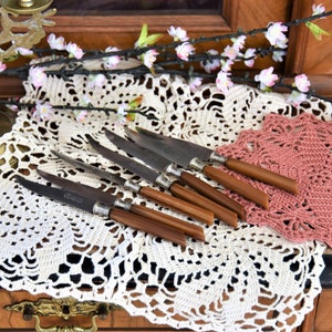 Könnte beinhalten: Ein Set aus sechs Vintage-Silbermessern mit Holzgriffen, angeordnet auf einer weißen Häkeldeckchen mit einem rosa Häkeldeckchen darüber.