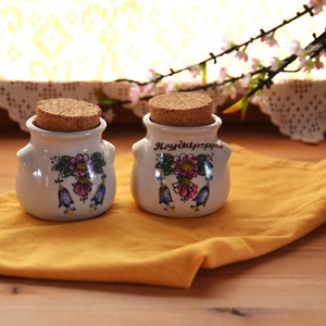 Könnte beinhalten: Zwei kleine weiße Keramikgläser mit Korkdeckeln und Blumendesigns. Die Gläser sind mit "Kryddpeppar" beschriftet und haben einen gelben Hintergrund. Die Gläser stehen auf einem gelben Tuch.