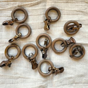 Könnte beinhalten: Eine Sammlung von Gardinenringen aus Holz mit Metallclips. Die Ringe sind in einem warmen Braunton gehalten, und die Clips haben eine Vintage-Messing-Oberfläche. Die Anordnung deutet auf ein Set von Gardinenbeschlägen hin.
