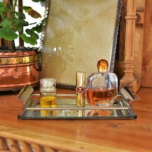 Könnte beinhalten: Ein silbernes Tablett mit einer durchsichtigen Glasflasche Parfüm, einem goldenen Lippenstift und einer durchsichtigen Glasflasche Öl. Das Tablett befindet sich auf einer Holzoberfläche.