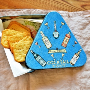 May include: A blue triangular tin with a cocktail theme. The tin has images of cocktails and bottles of liquor. The text on the tin reads "Huntley & Palmers Cocktail Biscuits". The tin is open and there are several biscuits inside.