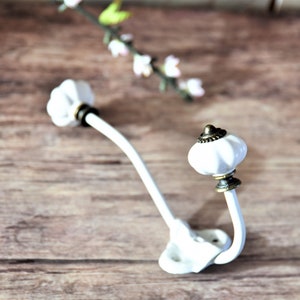 Pode incluir: Gancho de parede de cerâmica branca com um topo decorativo e arredondado e um braço curvo. O gancho tem uma estética vintage com detalhes em tom bronze. O gancho é montado numa base branca com dois orifícios para parafusos. Fundo de madeira.