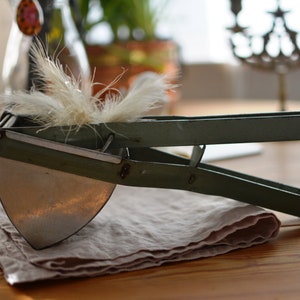 May include: An antique olive green potato ricer with a semi-circular blade and long handles. The ricer is resting on a folded beige linen napkin. A cluster of fluffy, light-colored feathers sits atop the ricer.