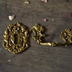 May include: Two ornate gold-toned metal door hardware pieces. The first piece is a keyhole cover with a decorative design featuring cherubs. The second piece is a handle with a similar decorative design.