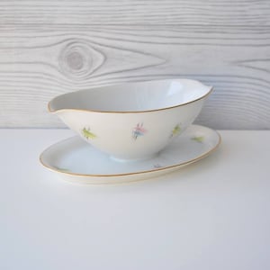 Könnte beinhalten: Eine weiße Porzellan-Sauciere und eine passende ovale Untertasse. Das Set hat einen Goldrand und ist mit farbenfrohen abstrakten Designs in Grün, Rosa und Blau verziert. Die Sauciere hat einen Ausguss.