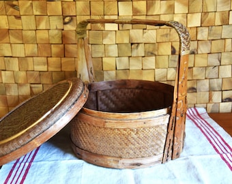 May include: A woven, brown, round basket with a lid and a handle. The basket is made of natural materials and has a rustic look.