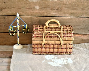May include: A woven wicker picnic basket with a red and brown pattern and a wooden handle. The basket is sitting on a beige fabric surface with a white floral design.