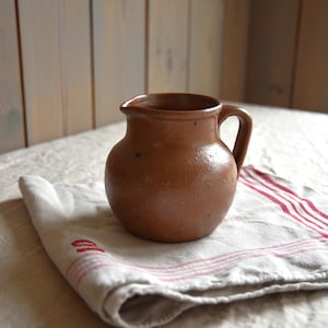 Könnte beinhalten: Ein kleiner, brauner, irdenen Krug mit Henkel. Der Krug steht auf einer weißen Leinen-Serviette mit roten Streifen.