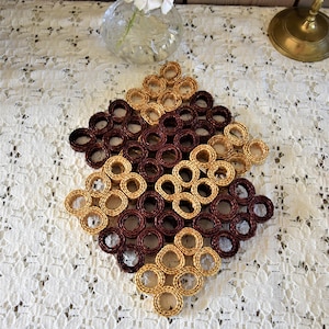 May include: A woven trivet in a diamond shape, featuring alternating sections of light tan and dark brown coiled material. The trivet is designed to protect surfaces from hot dishes and adds a rustic touch to any kitchen or dining area.