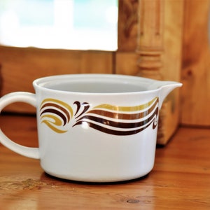 May include: White ceramic pitcher with a brown and gold geometric design. The pitcher has a handle and a spout.