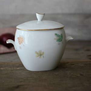 May include: White porcelain sugar bowl with a lid and two handles. The bowl has a gold trim and a floral design with green, yellow, and orange leaves.