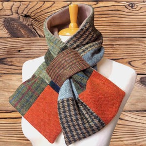 May include: A patchwork scarf constructed from various wool-like fabric panels featuring diverse patterns including herringbone, houndstooth, and plaid in shades of orange, green, brown, grey, and blue, draped over a light-coloured display form.