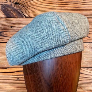 May include: A light gray flat cap featuring a textured herringbone pattern woven throughout the fabric. The cap has a structured, rounded crown and a short, stiff brim that sits flush against the main body of the hat, displaying a consistent weave.