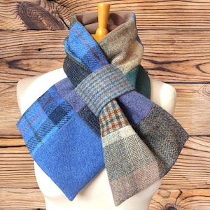 May include: A patchwork scarf featuring a variety of wool-like fabric patterns including blue plaid, brown herringbone, and grey tweed. The scarf is constructed from multiple rectangular sections of different textures and colours, joined together in a layered design.