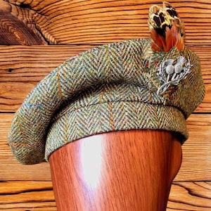 Può includere: Un berretto tam o' shanter in tweed a spina di pesce verde e marrone, caratterizzato da una spilla in metallo argentato a forma di cardo sul lato. Il cappello è decorato con un gruppo di piume naturali marroni e arancioni dietro l'emblema.