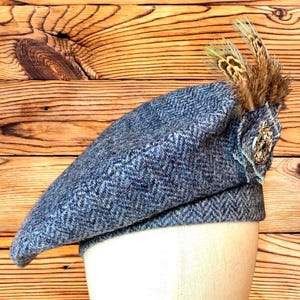 Può includere: Un basco blu in tweed con motivo a spina di pesce, ornato da piume marroni e gialle e da un emblema dorato. Il cappello è esposto su una testa di manichino su sfondo in legno.