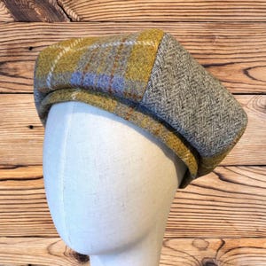 May include: A tweed flat cap with a plaid pattern in shades of yellow, blue, and brown, and a gray herringbone pattern. The cap is displayed on a mannequin head, showcasing its shape and design. The hat is a classic accessory.