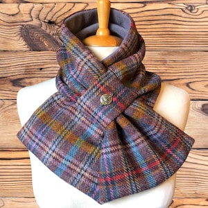 May include: A plaid scarf in shades of purple, blue, orange, and red, with a decorative button closure. The scarf is displayed on a mannequin, showcasing its design and texture. The scarf is made of a soft, warm material.