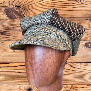 May include: A newsboy cap featuring a multi-colored herringbone pattern in shades of olive green, tan, and blue, with a distinct dark brown and tan textured panel on the side. The cap has a structured brim and a multi-paneled crown construction.