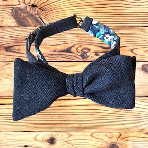Pode incluir: Uma gravata borboleta cinza escuro com forro estampado floral. A gravata borboleta é feita de um tecido texturizado e tem uma alça ajustável. O forro interno apresenta um design floral azul e branco.