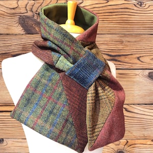 May include: A multi-coloured scarf featuring a patchwork of tweed fabrics in shades of green, brown, blue, and rust red. The scarf has a solid olive green lining and is secured with a square fabric loop, displaying a variety of plaid and herringbone patterns.