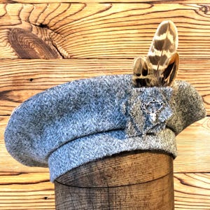 Può includere: Un basco di lana grigia con un emblema decorativo argentato e un gruppo di piume marroni e beige. Il basco è esposto su una superficie di legno, mettendo in risalto la sua consistenza e il suo design.
