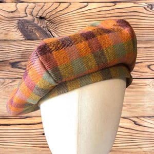 May include: A colorful plaid beret in shades of orange, green, pink, and brown. The hat is displayed on a white mannequin head against a wooden background. The beret has a classic shape and is made of a textured fabric.