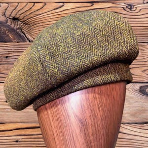 May include: A beret crafted from a textured wool fabric featuring a brown and mustard yellow herringbone pattern. The hat has a rounded crown and a structured band, displaying a dense weave with visible fibers throughout the material.