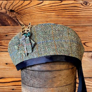 Può includere: Un basco in tweed verde e marrone con motivo a spina di pesce e sottili linee a quadri arancioni e blu. Il cappello è arricchito da una piccola tasca laterale contenente una piuma decorativa marrone e bianca e una spilla in metallo argentato.