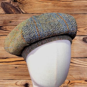 May include: A flat cap featuring a multi-colored tweed pattern in shades of olive green, brown, and blue. The fabric displays a classic herringbone weave with subtle orange and blue overcheck lines throughout the textured wool-like material.