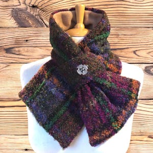 May include: A multi-coloured plaid scarf with a silver brooch. The scarf has shades of purple, green, orange, and brown. The scarf is displayed on a white mannequin, set against a wooden background.