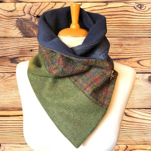 May include: A cowl neck scarf featuring a patchwork design of solid olive green wool, a multi-coloured plaid pattern with red and blue lines, and a dark blue herringbone fabric. The interior is lined with a solid dark blue fleece material.